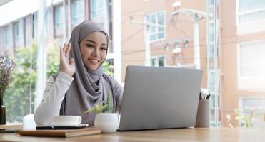 smiling beautiful muslim businesswoman using a laptop for online video meeting sitting at the desk in modern office space, looks at the webcam and waving hello photo