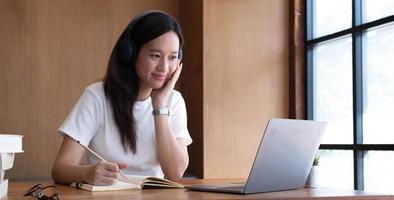 una estudiante sonriente usa audífonos inalámbricos para estudiar en línea con un maestro, una joven asiática feliz aprende el idioma escucha una conferencia mira un seminario web escribe notas mira una laptop sentada en un café, educación distante