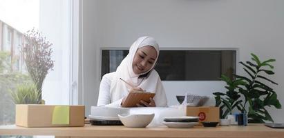 Religious asian muslim woman sitting and using smart mobile phone and talking to customer with happy face and business package SME box delivery background. photo
