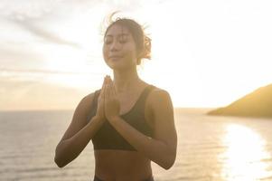 joven mujer asiática en ropa deportiva haciendo yoga en la roca en la playa durante el concepto de puesta de sol, salud y meditación foto