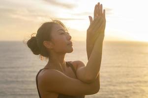 joven mujer asiática en ropa deportiva haciendo yoga en la roca en la playa durante el concepto de puesta de sol, salud y meditación foto