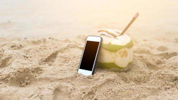 Fresh coconut with drinking straw with smartphone on the beach, travel and holidays concept. photo