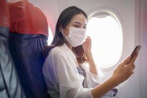 mujer joven con mascarilla está usando un teléfono inteligente a bordo, nuevo viaje normal después del concepto de pandemia covid-19 foto