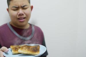 niño asiático muestra un plato de pan asado a la parrilla quemado con cara de mal humor infeliz - concepto de decepción de desayuno de comida foto