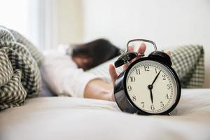 Sleepy woman reaching  holding the alarm clock in the morning with late wake up - every day life at home concept