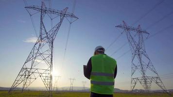 ingénieur électricien travaillant devant des lignes à haute tension. un ingénieur travaillant sur une tablette dans le domaine des lignes à haute tension. ingénieur électricien faisant une inspection sur le terrain. video