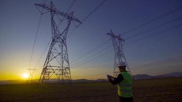 engenheiro trabalhando em frente a linhas de energia ao pôr do sol. um engenheiro com um tablet está trabalhando em frente a linhas de energia. realiza pesquisas de campo e investigações. video