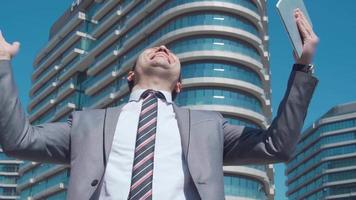 hombre de negocios alegría de éxito. el hombre de negocios se regocija trabajando en una tableta frente a edificios comerciales de lujo. Hombre de negocios exitoso video