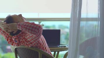 hombre de negocios sentado y descansando en el balcón del hotel. hombre sentado en el balcón con vista al mar, estirándose, relajándose y descansando. trabajando desde la computadora portátil. video