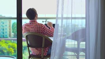 hombre de negocios sentado en el balcón del hotel. hombre de negocios sentado en la habitación del hotel con vista al mar. tiene una computadora portátil frente a él y está feliz. video