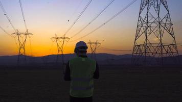 Engineer working in front of power lines at sunset. Engineer working on laptop at sunset and looking at electric poles. Field research and review video