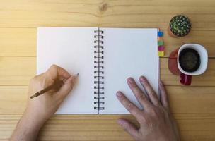 Top view of man writing note book on wooden table with coffee cup and small cactus pot photo