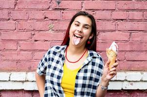 cheerful trendy woman with red hair eating ice cream on pink brick wall background at street photo