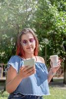 cheerful trendy woman with red hair drinking coffee at park, taking selfie photo