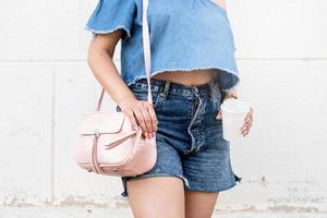 closeup of stylish woman with summer look, wearing jeans shorts, pink bag and holding cup of coffee photo