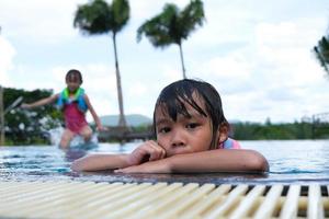 las niñas disfrutan nadando en la piscina. linda chica asiática que lleva un chaleco salvavidas se divierte jugando en la piscina al aire libre. actividades saludables de verano para niños. foto