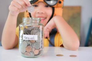 Cute little girl in a pilot hat is putting coins in a clear jar on the table to save for travel. Childhood dream imagination and Travel concepts. photo