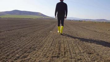 Bauer, der auf dem Feld spazieren geht. Der moderne Bauer, der eine Tablette hält, geht in gelben Stiefeln auf einem schmutzigen Ackerland spazieren. video