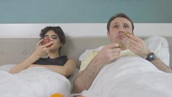 plaisir des fruits et de la télévision, un couple dans la chambre. le couple avec des fruits dans les mains regarde la télé dans sa chambre. video