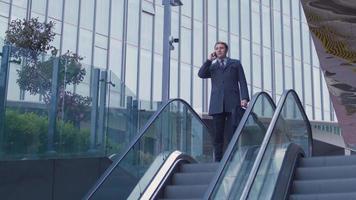 hombre de negocios bajando escaleras mecánicas, llamada telefónica. hombre de negocios bajando la escalera mecánica frente a un edificio de lujo. por un lado, está hablando por teléfono. video