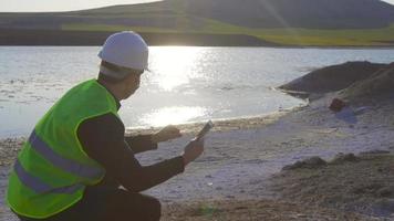 milieu en watervervuiling, ingenieur werken. de chemische vervuiling in het meer analyserend, werkt de ingenieur aan de tablet. er is wit chemisch afval bij het meer. video