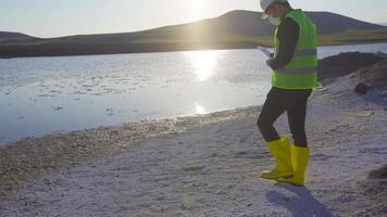 daño químico en el lago, contaminación ambiental. analizando la contaminación química en el lago, el ingeniero está trabajando en la tableta. hay desechos químicos blancos junto al lago. video