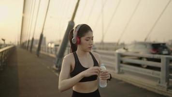 le donne atletiche asiatiche al rallentatore indossano abbigliamento sportivo per l'allenamento di corsa e si prendono una pausa per bere acqua in una bottiglia ogni giorno al mattino in una città con un fiume prima di andare alla competizione. video