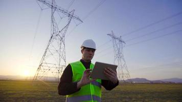 ingénieur travaillant devant les lignes électriques au coucher du soleil. un ingénieur avec une tablette travaille devant des lignes électriques. effectue des recherches et des enquêtes sur le terrain. video