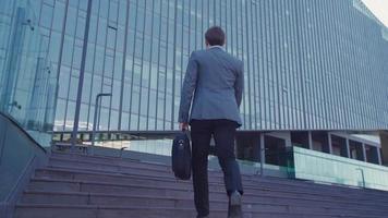 empresario con maletín caminando hacia el edificio. un hombre de negocios con traje que lleva un maletín camina hacia el edificio comercial. video