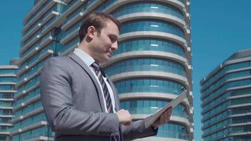 hombre de negocios que trabaja frente a edificios de lujo. un hombre de negocios con traje está trabajando en una tableta frente a edificios comerciales de lujo y un hombre de negocios está hablando por teléfono. video