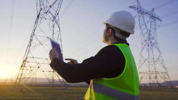 Ingenieur, der bei Sonnenuntergang vor Stromleitungen arbeitet. Ein Ingenieur mit einem Tablet arbeitet vor Stromleitungen. führt Feldforschungen und Untersuchungen durch. video