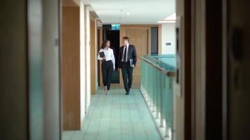 Businesswoman and businessman walking in the hallway. Businessman and businesswoman holding laptop are walking down the hall chatting. video
