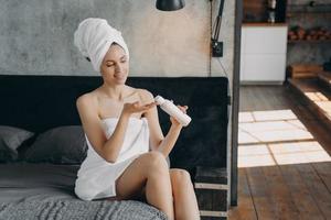 Happy girl applying body moisturizing lotion squeezing it out from bottle after bathing at home. photo