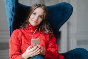 People and technology concept. Satisfied female meloman enjoys favourite music, listens audio record, dressed in red sweater, surfes in playlist, sits in armchair in living room, has spare time photo