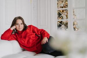 Indoor shot of pretty dark haired woman in warm winter clothes, leans at hand, lies on comfortable white sofa, looks directly at camera with calm expression. Domestic atmosphere. Time at home photo