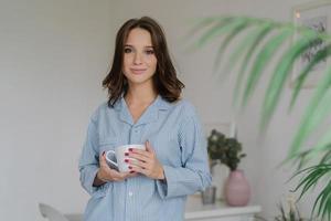 Morning time concept. Young pretty European woman in nightclothes carries white mug of coffee or cappuccino, stands indoor in living room, has weekend, enjoys good rest. People and lifestyle photo