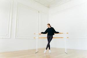 el bailarín de ballet clásico tiene ejercicios de estiramiento, se para cerca de las barras de ballet, usa sudadera negra y polainas, hace ejercicios en el estudio, se enfoca hacia abajo. instructora certificada de coreografía foto