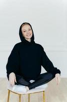 Relaxed cheerful woman in black sweatshirt, sits in lotus pose on chair, sits indoor, has glad expression, poses indoor, satisfied after active training with coach. People, rest, motivation concept photo