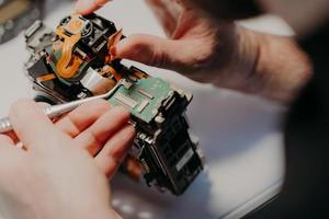 Hands of handyman repairing broken digital camera, holds special tweezers, works in workshop. Maintenance, occupation and repairing concept. photo