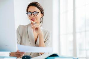Female economist poses in coworking space, focused in monitor, verifies or checks figures from financial report, holds paper document and pen, has dark combed hair, wears formal business suit photo