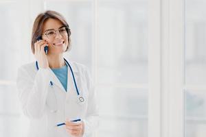 Shot of pleased female physician or surgeon gives prescription via mobile phone, has conversation with patient, holds pen, wears white uniform, works in hospital, stands indoor against window photo