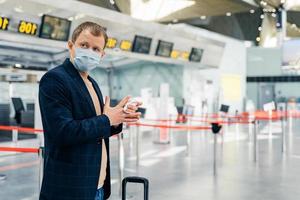 Photo of man passenger poses in airport, wears medical mask, uses antiseptic napkin to prevent spreading bacteria and coronavirus, cares about antibacterial treatment in public crowded place