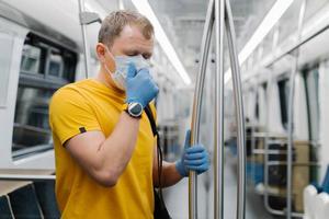 Horizontal shot of man passenger coughs and has problems with breathing, wears disposable mask and gloves, stands in public transport, metro, prevents from coronavirus. Public health solution photo
