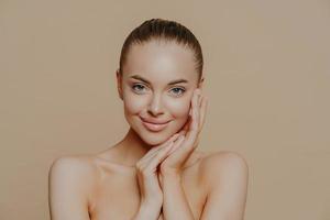 Natural beauty portrait. Photo of young woman enjoys perfect fresh skin after visiting cosmetologist, smiles gently, poses naked against beige background, touches face, wears minimal makeup.