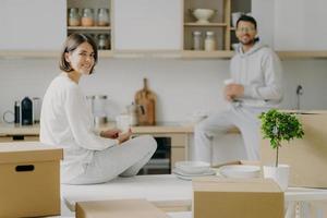 Horizontal shot of happy young woman and man sit on kitchen desk, drink coffee, have break after unpacking boxes, have pleasant talk, surrounded with plates, indoor plant in pot. Moving concept photo
