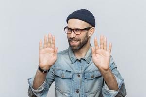 Dont bother me Cheerful male with pleasant appearance shows stop sign, asks not to speak with him, wears fashionable hat and glasses, isolated over grey studio background. People and denial concept photo