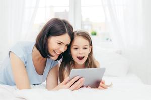 Positive cheerful small female kid and her young mother watch interesting cartoon on tablet, connected to wireless internet, pose against bedroom interior, have happy expressions. Family concept photo