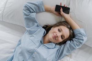 Relaxation concept. Pretty girl with pleased satisfied expression, dressed in pyjamas, rests in bed, holds smart phone, listens pleasant music in earphones. View from above. Woman has good day at home photo