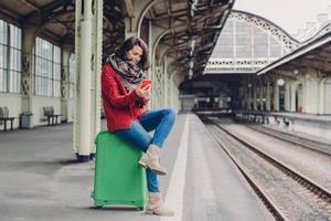 Pleased young woman has positive look, sits crossed legs at bag, holds cell phone, searches place on online map, makes video call, surfes internet website, waits for train at railway station photo