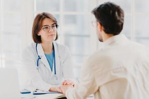 Friendly female doctor tries to support patient, holds his hands, gives useful consultation and explains medical information, makes diagnostic examining, pose in hospital room. Heathcare, assistance photo